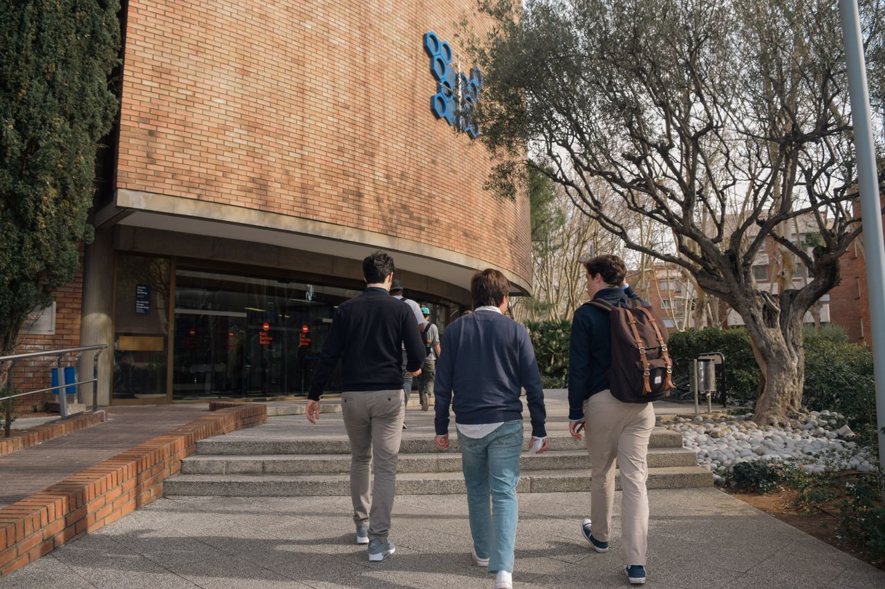 Three students approach a university building entrance in Barcelona, Spain on a sunny day.